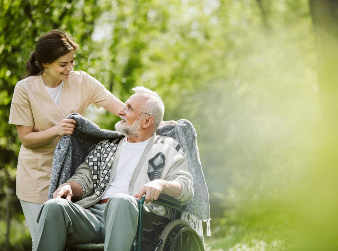 Carer wrapping their service user with a blanket standing outside while the service user is seated in a wheelchair.