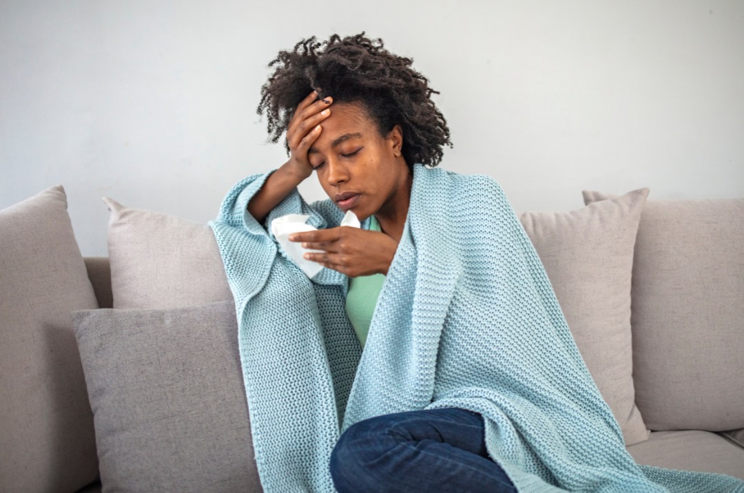 Woman sat in her living room, covered with a blue blanket. With a tired look on her face, hand on her head in exhaustion and a tissue in her other hand.