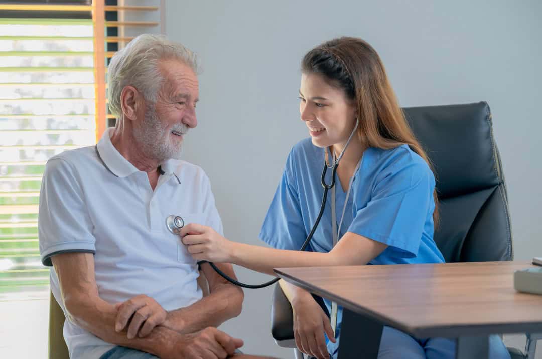 Nurse using a stethoscope on an elderly man, both are sat down in a well lit office.