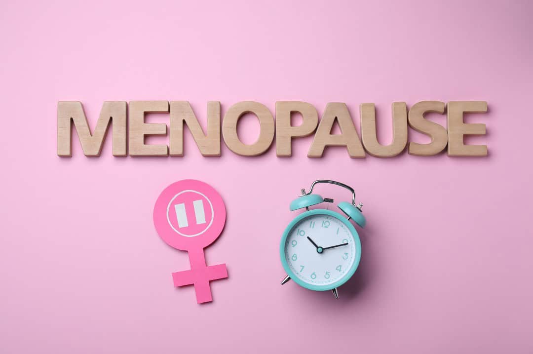 Pink background with the text 'Menopause' over the top. Below the text is a darker pink female symbol with a pause symbol in the centre of the circle. Next to this is a blue alarm clock.