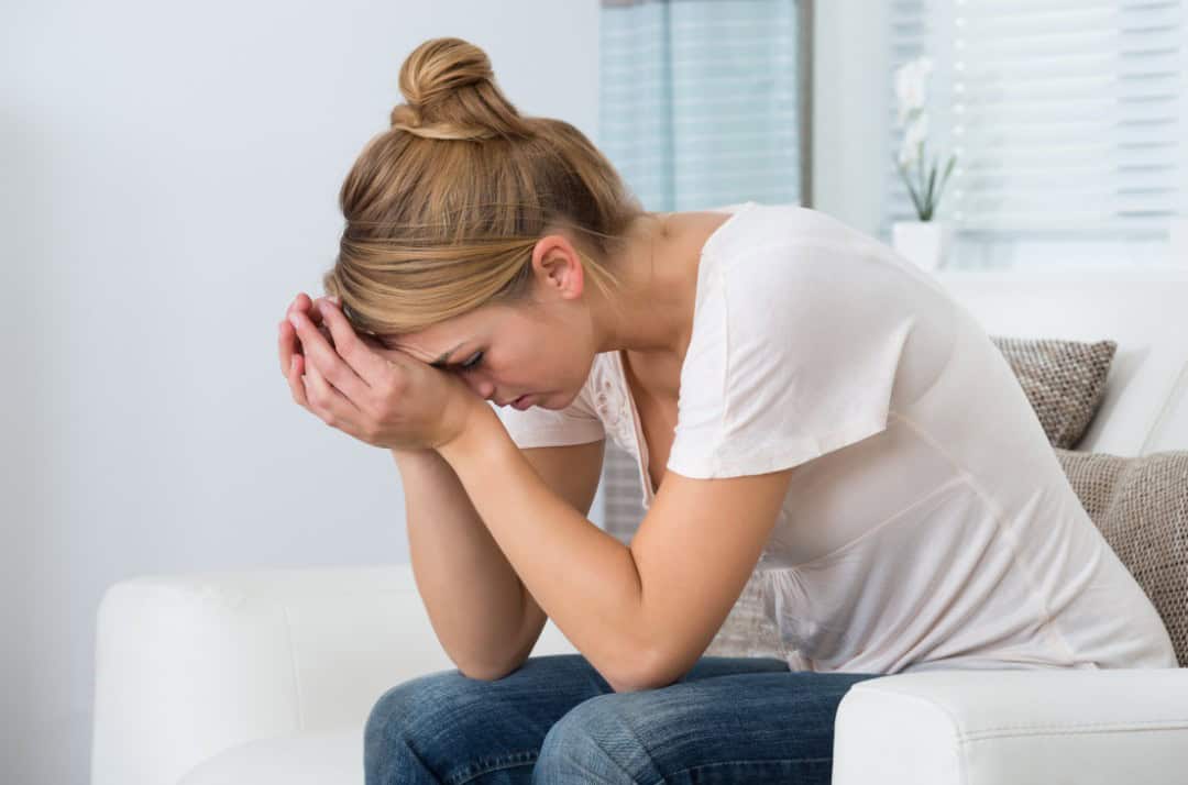 A woman with a look of stress on her face, sat down on a couch with her head on her hands.