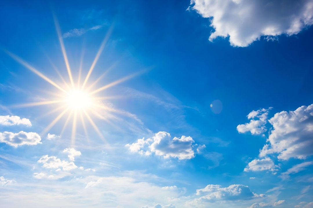 Imagine of an extremely bright sun among a blue sky surrounded by some white clouds.