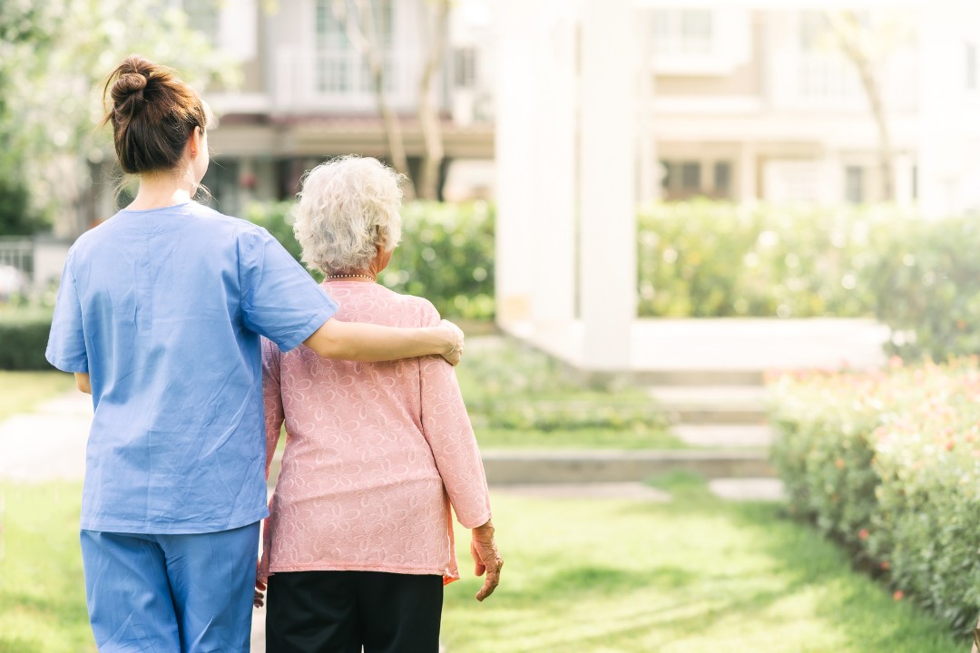 A carer supporting an older woman to walk outside in a garden on a sunny day.