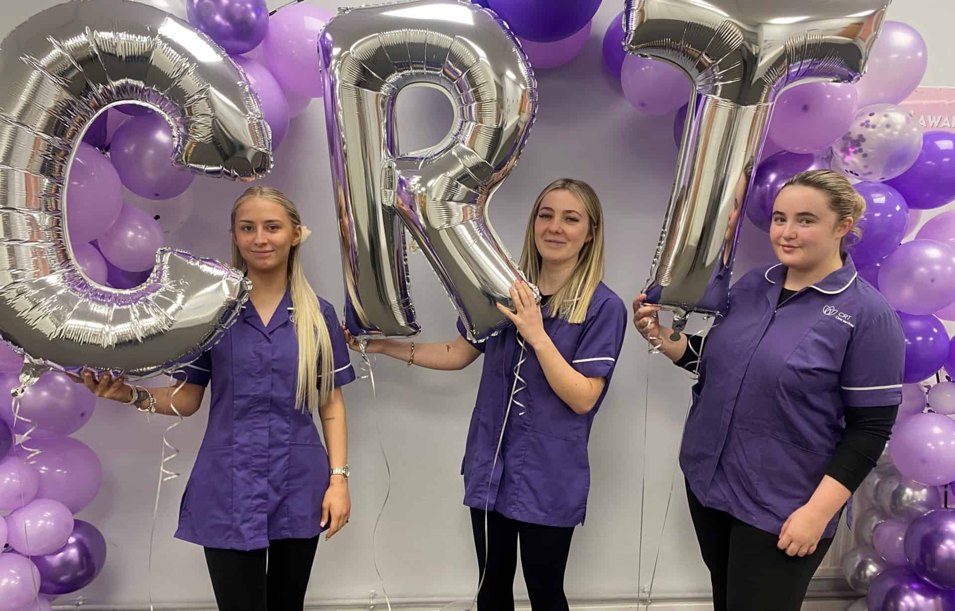 Three CRT Home Care Workers standing in front of a white wall with a purple balloon arch holding silver balloons that spell out 'CRT'.